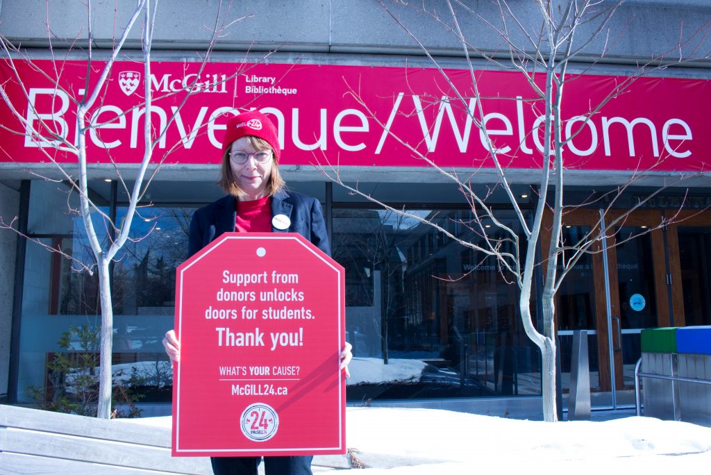 Dean Colleen Cook wearing a red McGill24 toque and t-shirt standing in front of the red Bienvenue/ Welcome banner holding a red poster that says "Support from donors open doors for students""