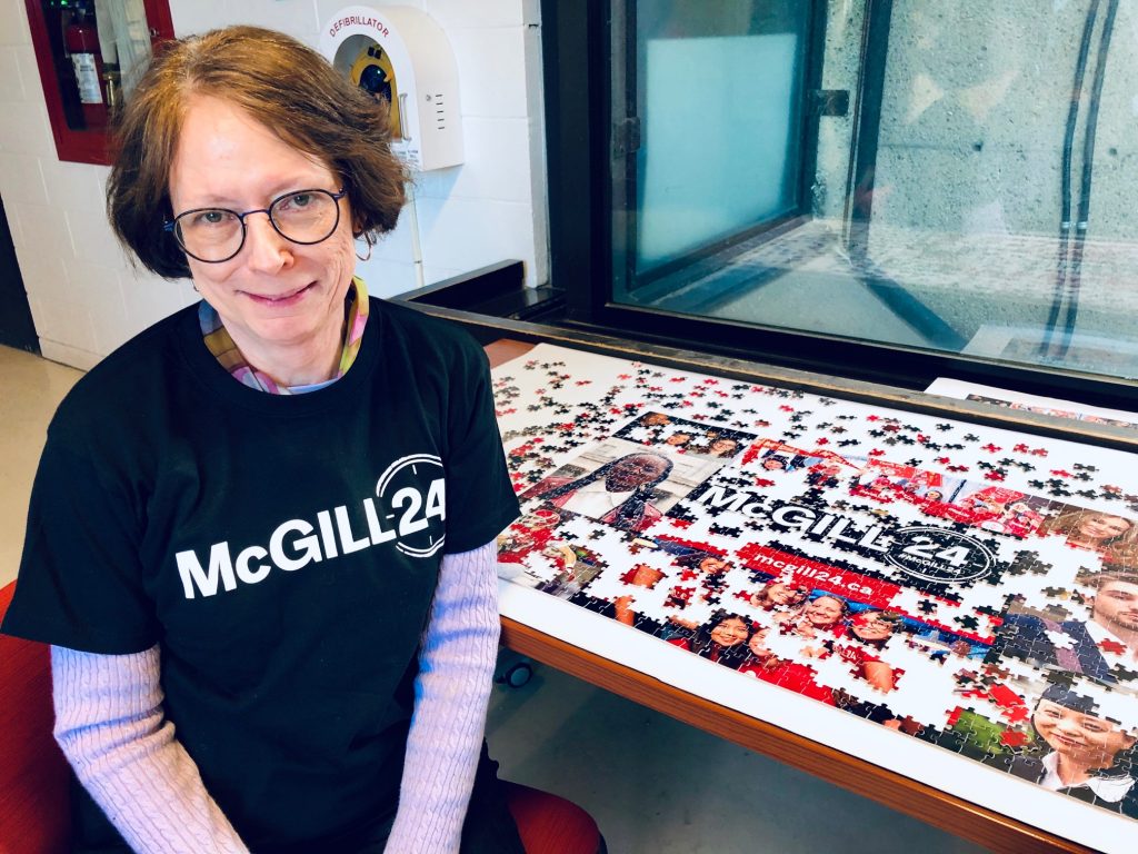 Dean Colleen Cook wearing a black McGill24 t-shirt sitting in front of a partially completed McGill24 puzzle featuring a collage of different photographs.