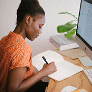 Woman sitting at a desk writing in a notebook with a file open on a desktop.