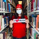 Student Alisa Nosova wearing a mask and a red McGill24 hat standing in Library stacks with a cut-out that says "J'aime McGill".