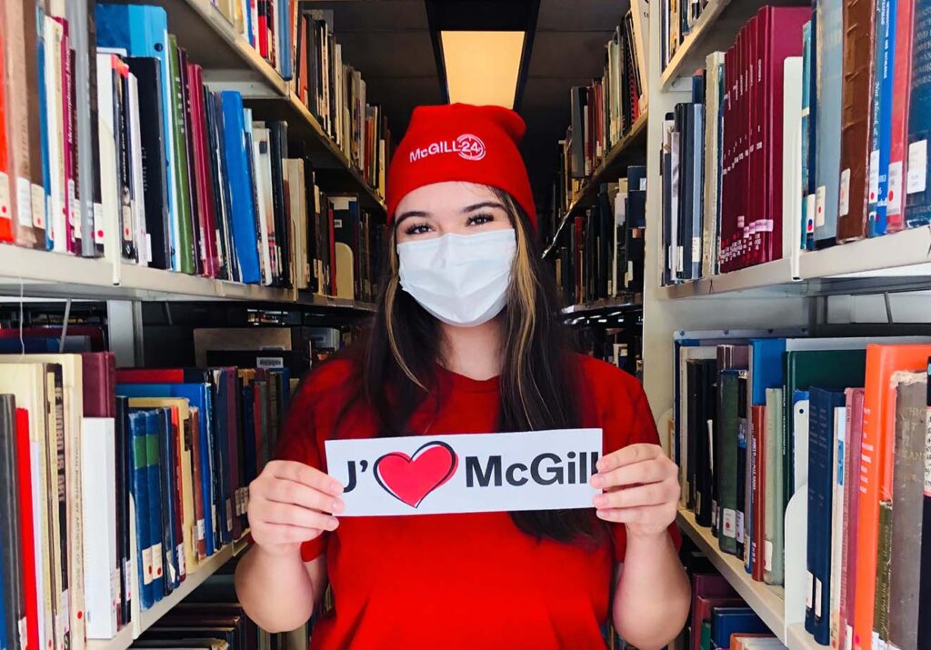 McGill student in red McGill24 hat in Library stacks holding up a sign that says "J'aime McGill".