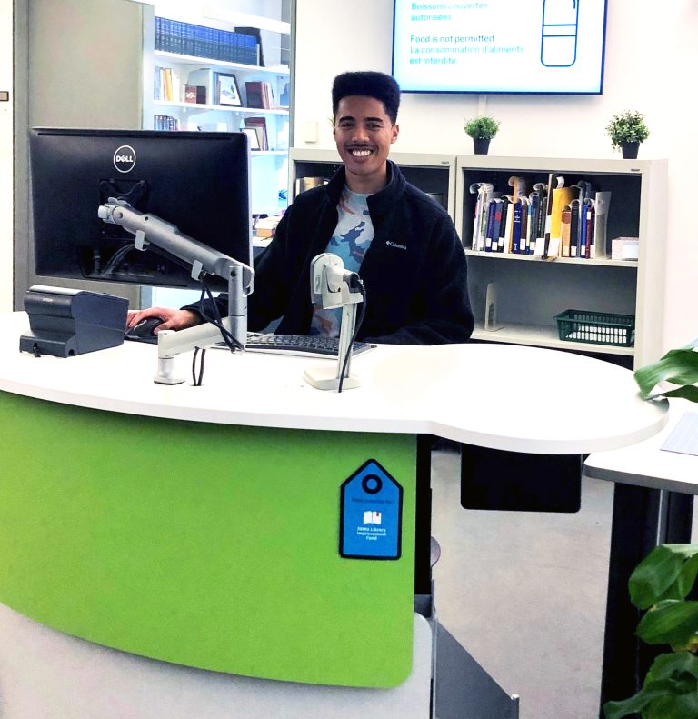 Alexander Norton III sitting at a desk in front of a computer in the Music Library smiling at the camera.