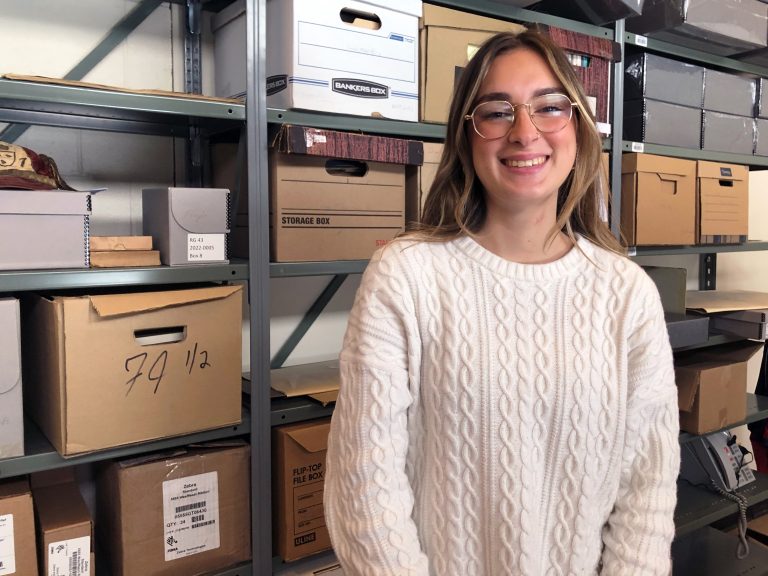 Emma Gallanti smiling at the camera standing in front of a shelving unit filled with boxes.