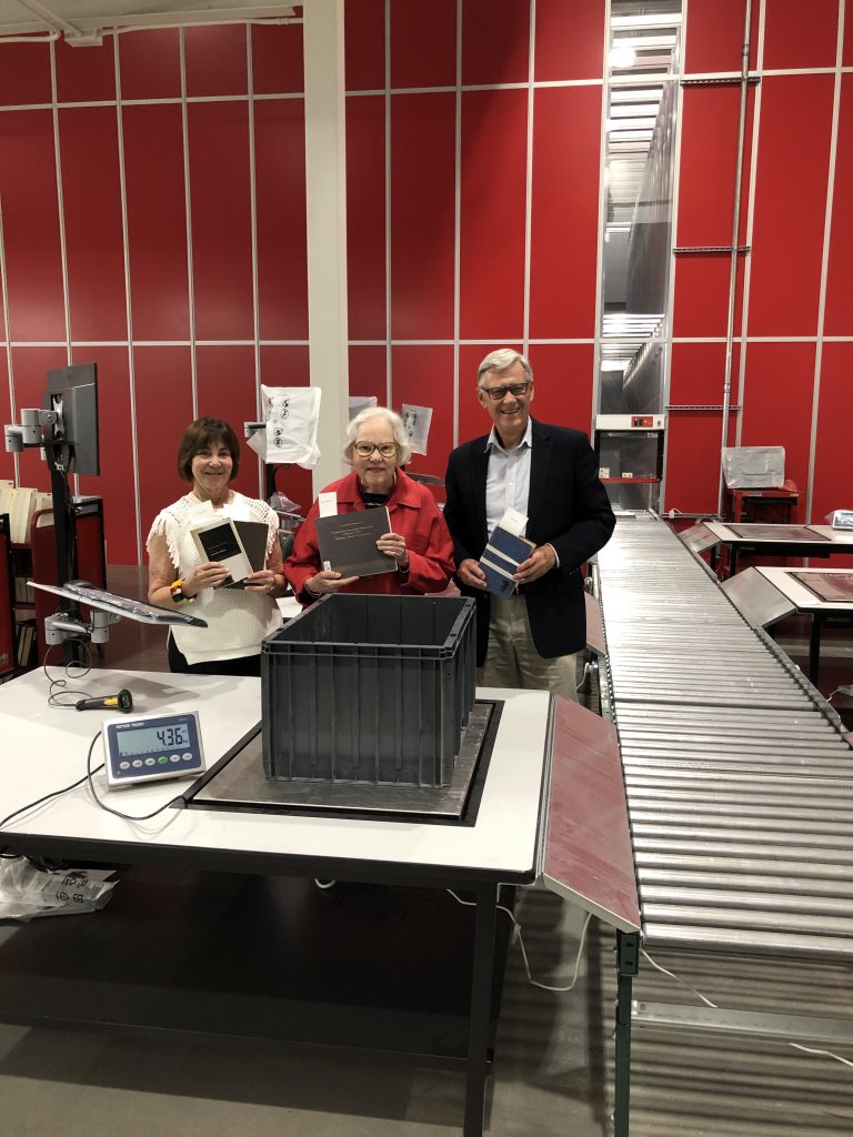 From left to right: Lenore Harris, Louise Robertson, and Don Walcot at the McGill University Collections Centre holding up books in front of red wall.