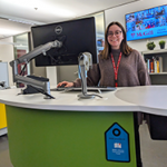 Person smiling at the camera from behind a green desk with a tag on it indicating student support.